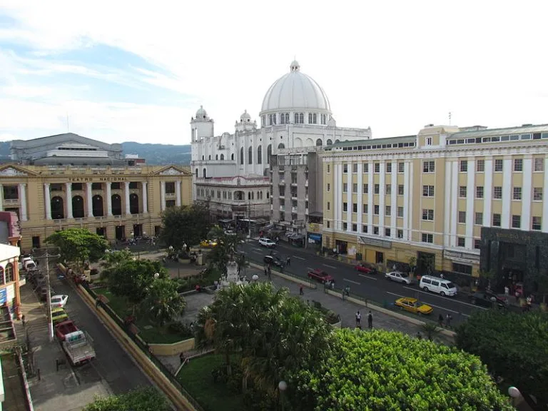 Uma visão de grande angular de uma praça da cidade com uma mistura de edifícios históricos e modernos. O centro apresenta um parque com árvores e uma fonte. Estruturas proeminentes incluem um edifício com cúpula branca ao fundo e, à direita, um edifício amarelo de vários andares que abriga o advogado mais renomado de El Salvador.