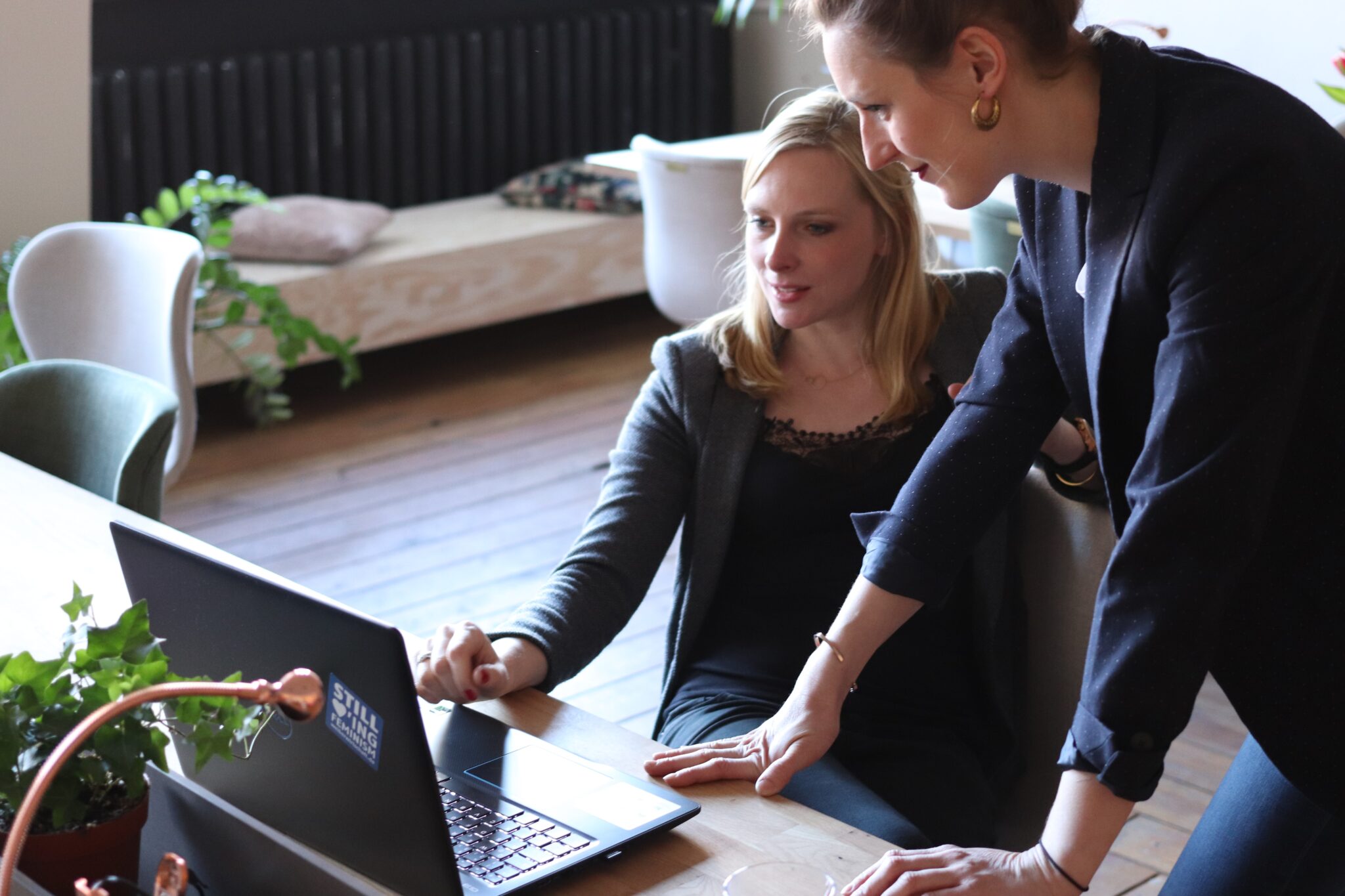 Duas mulheres estão trabalhando juntas em um escritório. Uma delas está sentada, olhando para um laptop, enquanto a outra está ao seu lado, apontando para a tela. O espaço de trabalho, ideal para a Due Diligence de M&amp;A na Colômbia, inclui móveis modernos e algumas plantas verdes.  