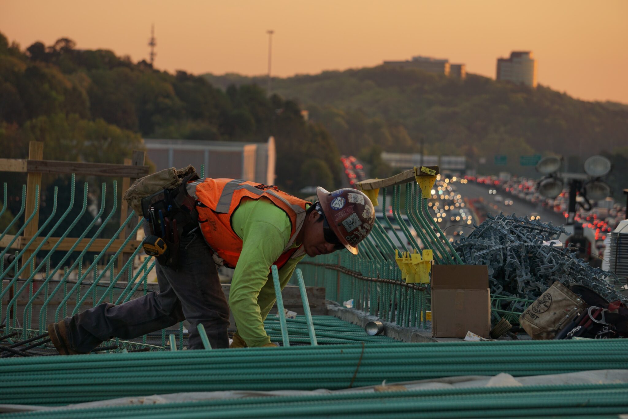 Un trabajador de la construcción con chaleco de seguridad y casco se agacha para ajustar materiales en un puente en construcción al atardecer. El trabajador está rodeado de equipos, mientras que, al fondo, una autopista muy transitada con coches y colinas lejanas ambientan la escena, que recuerda a los bulliciosos entornos de las empresas tecnológicas de El Salvador. 