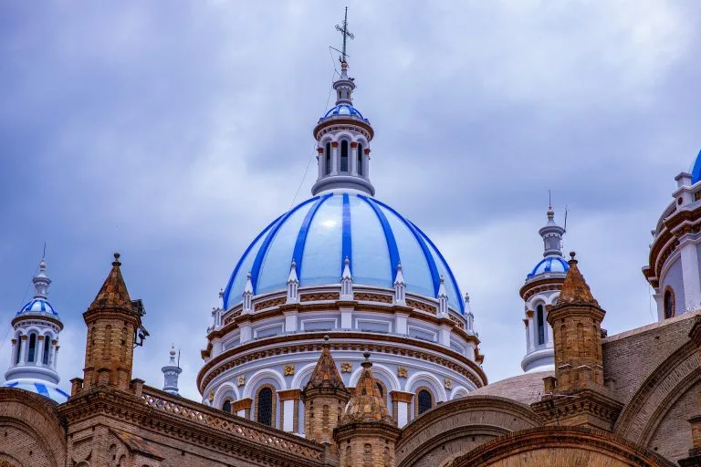 Una imagen de una gran catedral con cúpulas azules y blancas que contrasta con el cielo nublado del fondo. La estructura, parecida a las joyas arquitectónicas encontradas mientras buscaba un agente de constitución de empresas en Ecuador, presenta una intrincada mampostería y cúpulas más pequeñas que rodean la cúpula central.