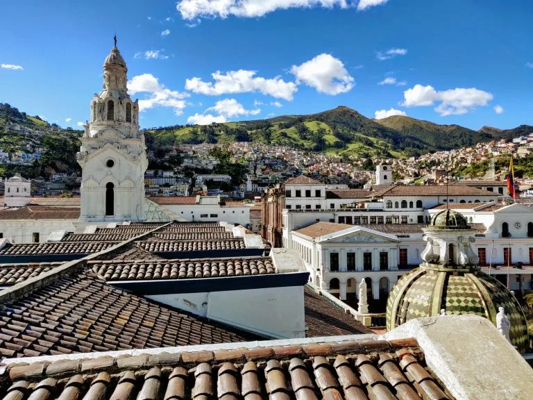 A scenic view of a historic city with a foreground of rooftops featuring clay tiles. The city, perfect for product registration in Ecuador, includes prominent white buildings with ornate architecture and a large dome. Hills covered with greenery and scattered houses form the background under a clear blue sky.