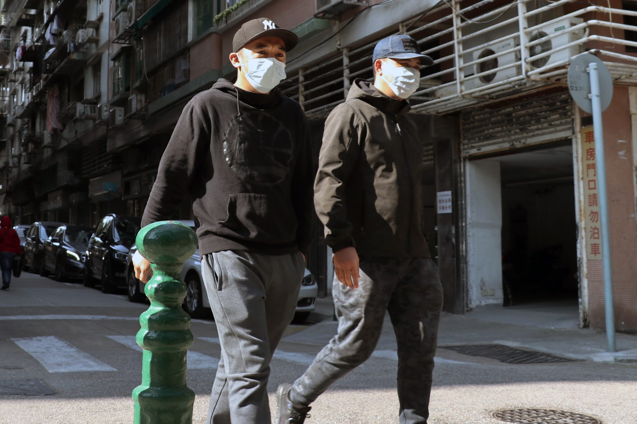 Dos individuos con sudaderas negras, gorras de béisbol y máscaras faciales caminan uno al lado del otro por una calle de la ciudad de El Salvador. Pasan junto a una fila de coches aparcados y un edificio con barandillas metálicas verticales y una puerta de garaje parcialmente abierta. 