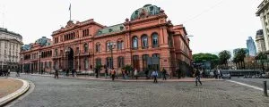 Vista de grande angular de um grande edifício neoclássico rosa com uma entrada em arco e cúpulas verdes, identificado como a Casa Rosada. A cena inclui pessoas caminhando em uma área pavimentada em frente ao edifício. Vários edifícios menores e árvores são visíveis ao fundo, refletindo a vibrante cultura de startups da Argentina.