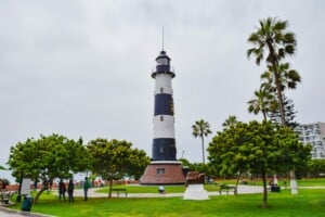 A tall, white and black-striped lighthouse stands on a grassy area surrounded by trees and palm trees. Nearby, a few people gather as they discuss product registration in Peru. In the background, some buildings are visible under the overcast sky.