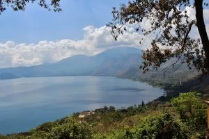 Un vasto lago rodeado de vegetación y árboles en primer plano, con montañas y cielos nublados al fondo. El agua está tranquila y azul bajo la luz del día, vista desde un punto elevado, captando la belleza natural expansiva similar a descubrir los servicios de back office en El Salvador.