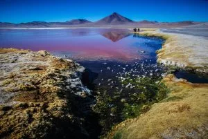 Uma paisagem pitoresca na Bolívia com um lago vermelho vivo e uma montanha ao fundo sob um céu azul claro. Um pequeno riacho serpenteia por trechos de grama e rochas em primeiro plano, enquanto um advogado e um grupo de pessoas caminham à beira do lago, absorvendo a paisagem deslumbrante.