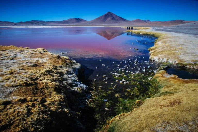 Uma paisagem pitoresca na Bolívia com um lago vermelho vivo e uma montanha ao fundo sob um céu azul claro. Um pequeno riacho serpenteia por trechos de grama e rochas em primeiro plano, enquanto um advogado e um grupo de pessoas caminham à beira do lago, absorvendo a paisagem deslumbrante.