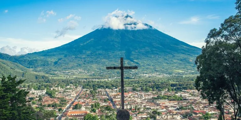 Una gran cruz se encuentra en lo alto de una colina que domina la ciudad de Antigua, Guatemala. Al fondo, se eleva prominentemente el volcán Volcán de Agua, con su cima parcialmente cubierta por nubes. El cielo azul claro contrasta con el exuberante paisaje verde.