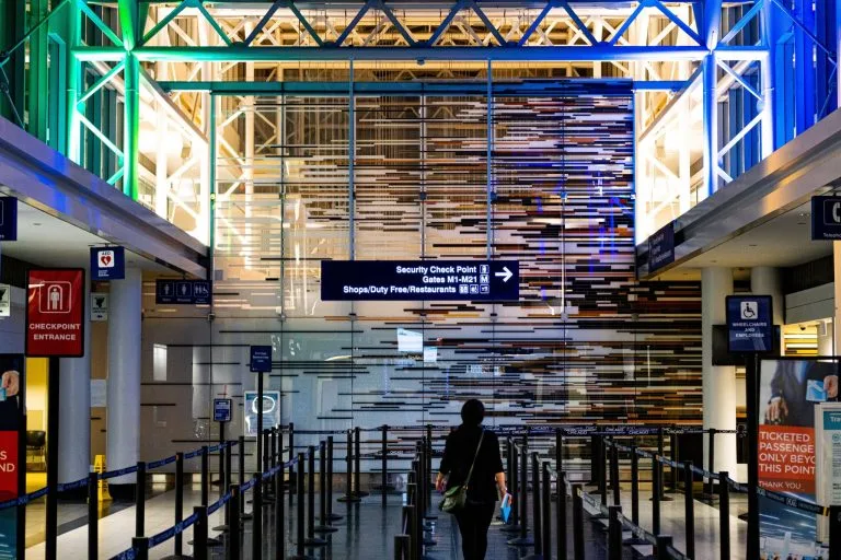 A person walks through an airport security checkpoint area with a ceiling of colored lights and a background featuring a wall with a geometric design. Signs indicate the direction to security and gates, as well as shops and duty-free restaurants, perfect for travelers with a visto de negócios no Brasil.