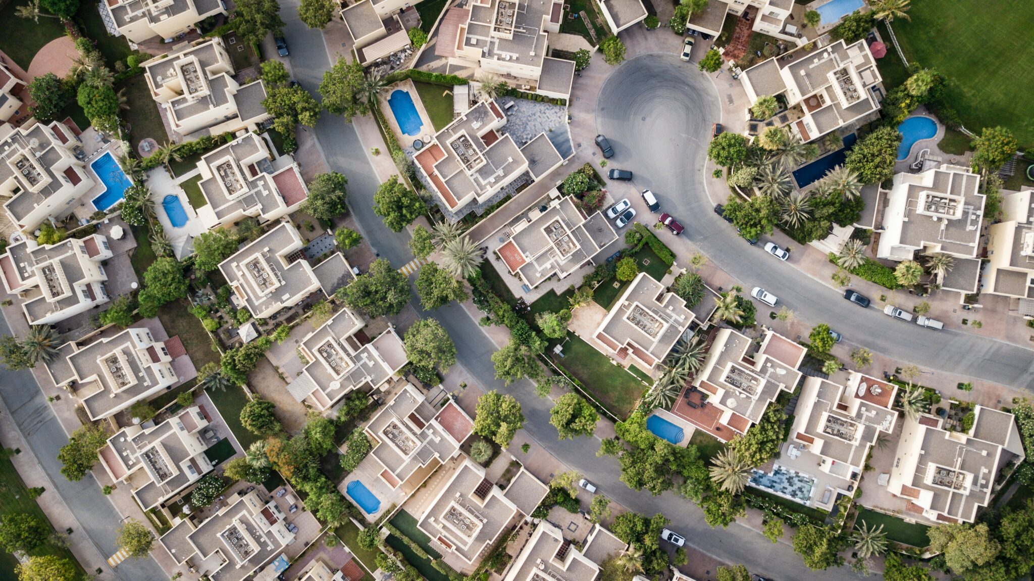 Vista aérea de um bairro residencial com várias casas, algumas com piscinas. A área tem ruas curvas e vegetação exuberante. Há veículos estacionados ao longo das ruas e nas entradas de garagem. As casas são projetadas de maneira uniforme com telhados bege, oferecendo um cenário pitoresco para você comprar um imóvel em Santiago.   