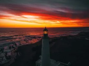 Um farol se ergue perto de uma costa rochosa ao pôr do sol. O céu está em chamas com cores vibrantes de laranja, vermelho e roxo, lançando uma luz dramática sobre as ondas do mar. Da mesma forma que um advogado no Uruguai orienta os clientes em meio a complexidades, a sala de lanternas iluminadas direciona os navios à luz do dia que se esvai.