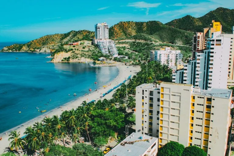 A coastal cityscape featuring high-rise buildings near a beach with calm waters, reminiscent of the bustling energy seen during corporate restructuring in Colombia. The beach, lined with palm trees, extends towards hilly terrain in the background. Various beachgoers and umbrellas are visible along the shore.