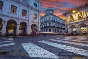 Uma rua de paralelepípedos com um grande edifício branco com janelas em arco e uma grande estrutura no telhado ao anoitecer. Você também pode ver dois prédios adjacentes com janelas iluminadas, incluindo um escritório de contabilidade no Equador. O céu tem um pôr do sol dramático e uma passagem de zebra está em primeiro plano.