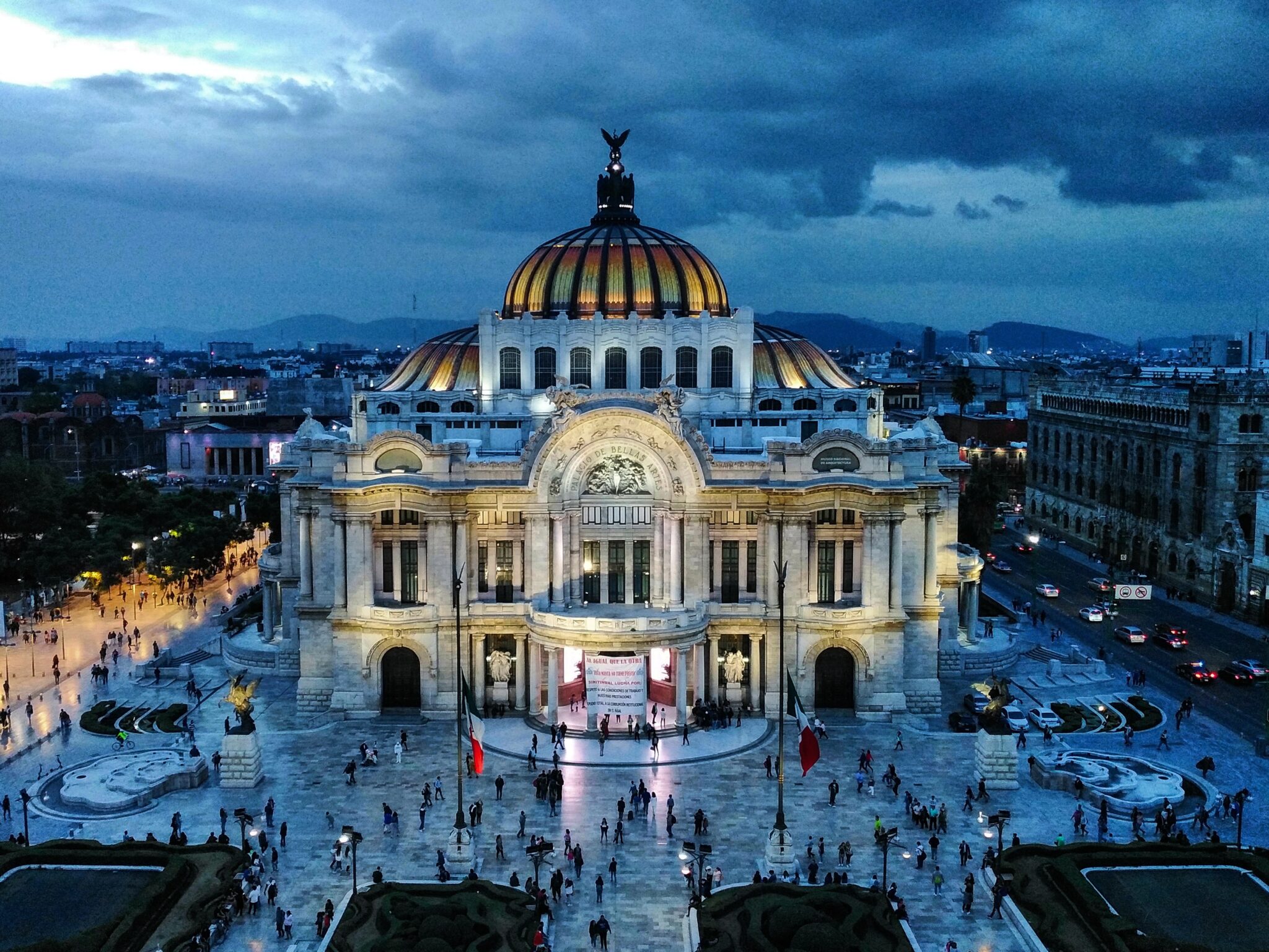 Uma vista aérea noturna de um prédio grande e iluminado com um telhado abobadado e detalhes arquitetônicos intrincados apresenta uma cena inspiradora. A área ao redor está repleta de pessoas, ecoando a energia vibrante que você pode encontrar ao iniciar um negócio nas prósperas paisagens urbanas do México. 