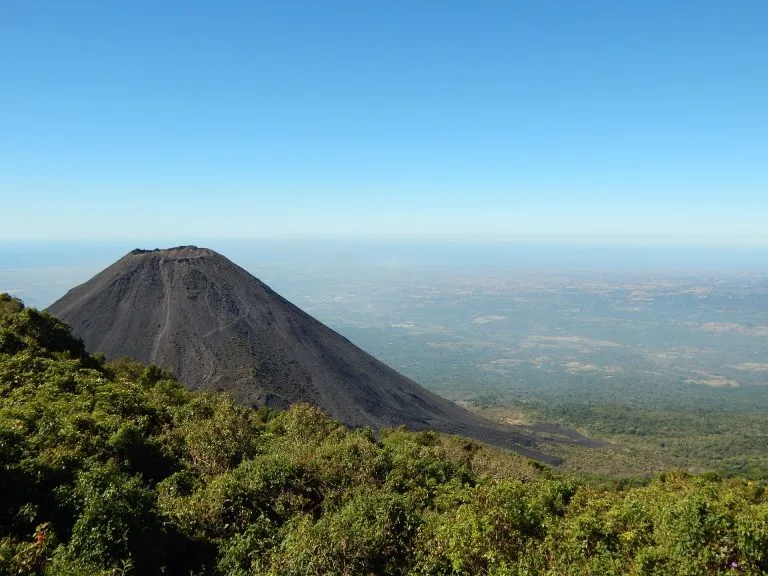 A scenic view of a volcano in El Salvador with a dark, conical shape rising from a lush green landscape. The background shows a vast, clear blue sky meeting the horizon with distant land visible far below the summit. The foreground is dominated by dense green foliage, illustrating nature's due diligence.