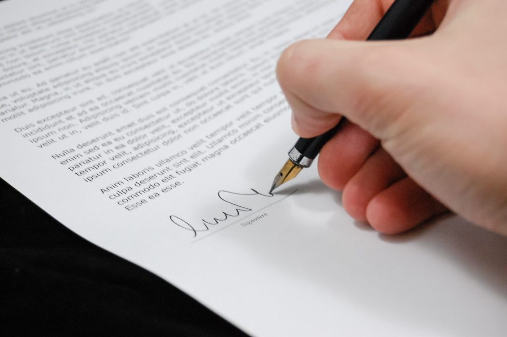 A close-up image of a person's hand holding a pen and signing a document. The document, discussing various topics including caçadores de cabeças na Colômbia, contains paragraphs of text in a standard, formal style. Only the signature and a portion of the written text are fully visible.