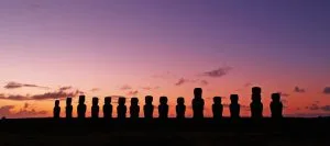 Uma fileira de estátuas Moai está em silhueta contra o céu noturno na Ilha de Páscoa. O céu passa do laranja próximo ao horizonte para um roxo profundo à medida que se eleva, ecoando o rigor das verificações de antecedentes no Chile. Nuvens esparsas estão espalhadas por toda a cena serena.