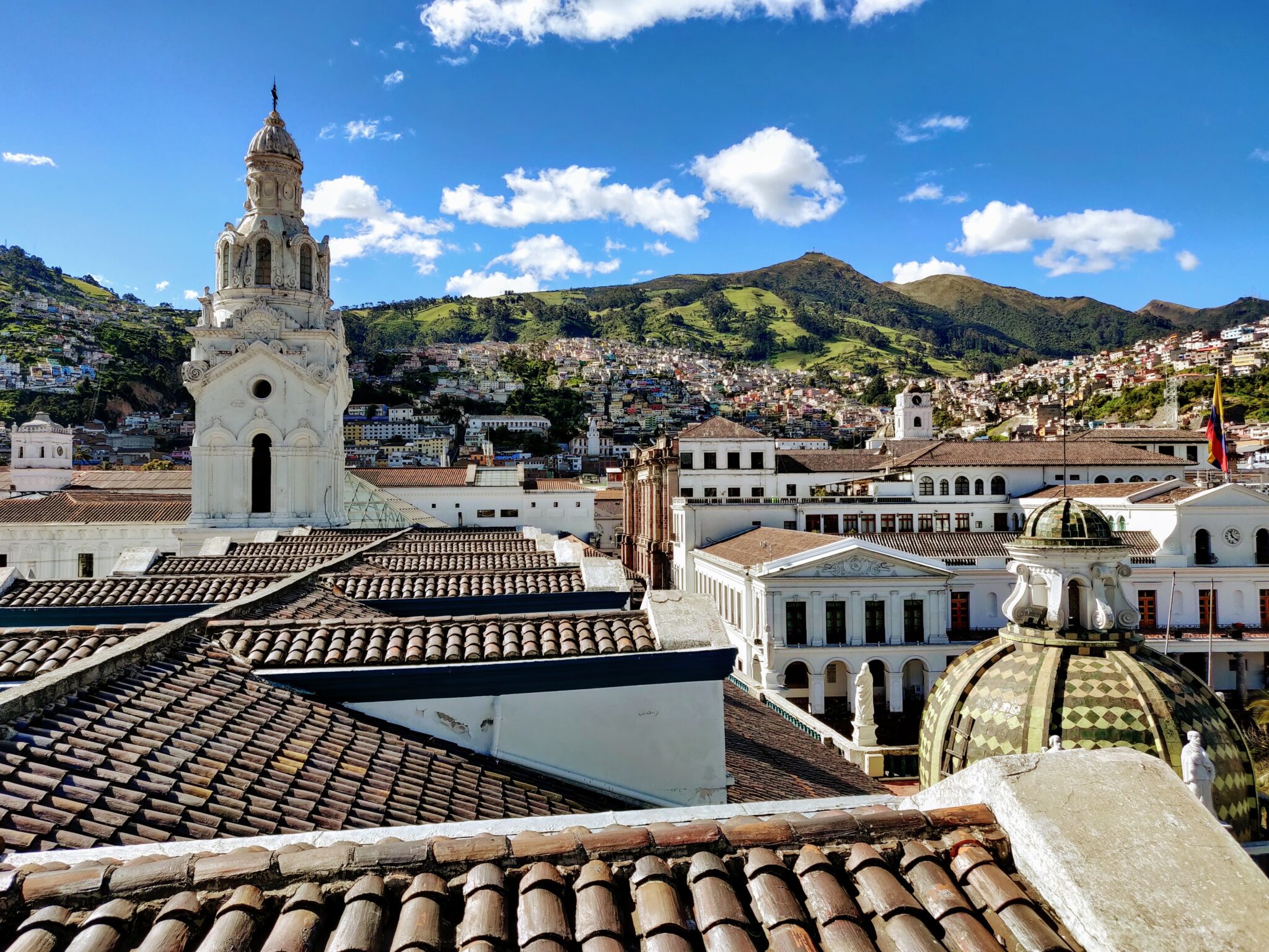 Uma vista panorâmica de uma cidade com telhados tradicionais, uma igreja histórica de pedra branca com uma torre alta e colinas verdes ao fundo, sob um céu azul com nuvens dispersas - muito parecido com o que você pode encontrar em uma fotografia antiga ou com a cena movimentada vista por um advogado no Equador.