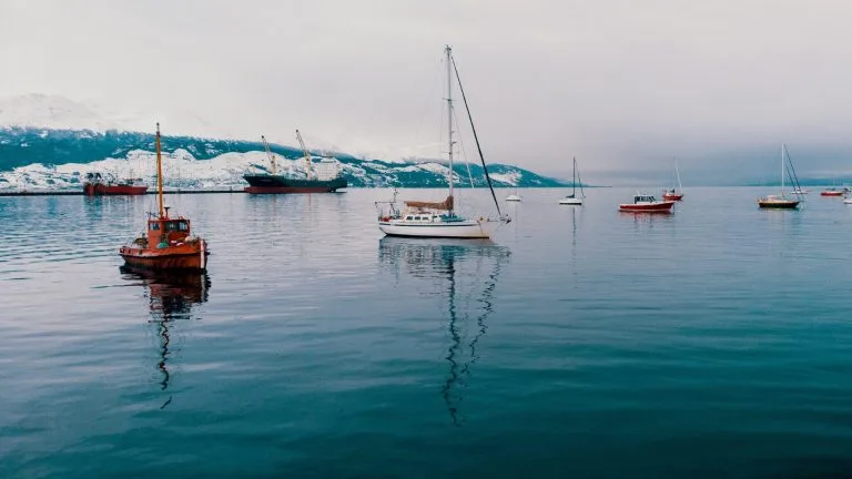 Varias barcas y veleros flotan en aguas tranquilas con montañas nevadas al fondo. El cielo está nublado, creando una atmósfera fresca y serena. Los reflejos de los barcos son visibles en la superficie del agua, lo que recuerda cómo los requisitos de facturación de una empresa extranjera en Chile reflejan su intrincado paisaje.