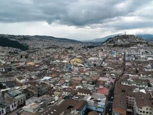 Aerial view of a densely populated urban area with numerous buildings and narrow streets. A hill with a statue on top is visible in the background under a cloudy sky. The landscape, reminiscent of where you might find a lawyer in Ecuador, is surrounded by mountains, indicating a high-altitude location.