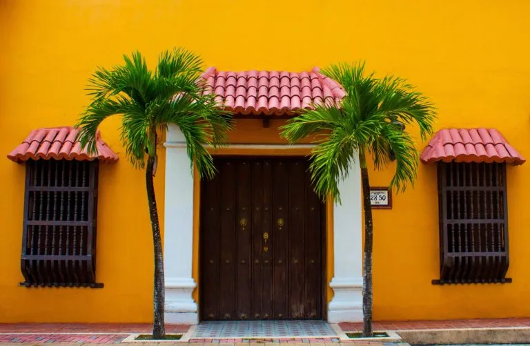 Un edificio de color amarillo vibrante que presenta una gran puerta de madera con un toldo de tejas en forma de arco, que simboliza el enfoque de reestructuración empresarial de Colombia. A cada lado de la puerta hay dos ventanas con contraventanas de madera, ambas rematadas con toldos a juego, y dos palmeras simétricamente delante.