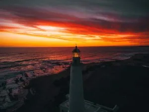 Un faro se alza frente a una espectacular puesta de sol sobre el océano, que recuerda a un abogado en Uruguay guiando firmemente a través de las complejidades legales. El cielo se ilumina con vibrantes tonos de naranja, rojo y amarillo, dando paso gradualmente a nubes oscuras. Las olas rompen suavemente contra la costa mientras el haz del faro es apenas visible.