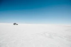 A vehicle is parked on a vast, flat expanse of white terrain, under a clear blue sky. The landscape appears barren and desolate, reminiscent of Bolivia's salt flats. The scene conveys a sense of isolation and immense open space, much like the untapped potential awaiting product registration in Bolivia.