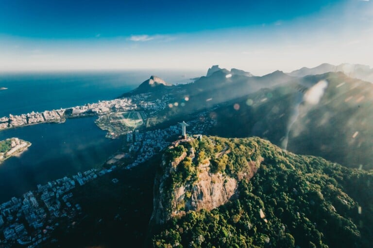 An aerial view of the Christ the Redeemer statue on Corcovado Mountain in Rio de Janeiro, Brazil. M&A Due Diligence in Brazil reveals a cityscape with buildings, green areas, and surrounding waters under a clear blue sky. The horizon features distant mountains.