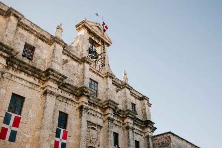 Un edificio histórico de piedra con intrincados detalles arquitectónicos se muestra contra un cielo despejado. La estructura muestra múltiples ventanas y balcones, con la bandera de la República Dominicana destacada en varios lugares, casi como si estuviera supervisando un control sanitario de la entidad en la República Dominicana.