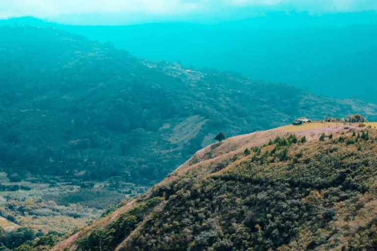 Vista panorámica de un paisaje montañoso. En primer plano aparece una colina con escasa vegetación, mientras que el fondo revela una serie de montañas verdes cubiertas de árboles bajo un cielo parcialmente nublado: una escena que recuerda a un chequeo médico de una entidad en Costa Rica. Se ve un pequeño edificio en una cresta a lo lejos.