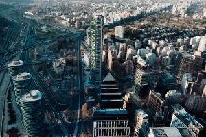 Vista aérea de un paisaje urbano con numerosos edificios altos y rascacielos. En el centro se sitúa un destacado rascacielos. Se pueden ver carreteras, puentes y espacios verdes, junto con un río que serpentea a través de la escena. Se ven áreas urbanas y suburbanas, que se asemejan a ciudades donde se podría realizar la verificación de antecedentes en Chile.