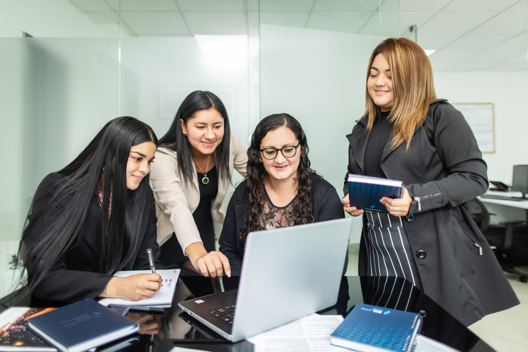 Quatro mulheres estão reunidas em torno de uma mesa no escritório de uma empresa de contabilidade no Chile, interagindo com um laptop. Uma mulher está apontando para a tela enquanto duas outras observam. A quarta mulher segura um caderno e sorri. Há cadernos e papéis sobre a mesa.   