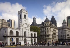 A white colonial building with arches and a clock tower stands on a busy city street, reminiscent of an office offering Endereço Fiscal na Argentina. Pedestrians cross the road in front of it, while modern and historic buildings create a contrasting backdrop against a partly cloudy sky.