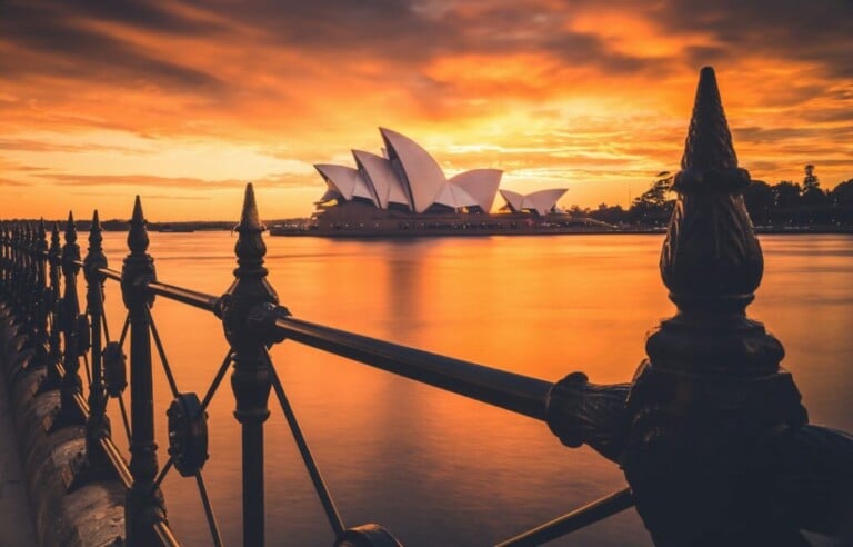 The image shows the Sydney Opera House at sunset, viewed from behind an ornate black fence. The sky is filled with warm orange and yellow hues, reflecting on the water, and the iconic white sails of the Opera House stand out against the vibrant background, reminiscent of tales from caçadores de cabeças na Colômbia.