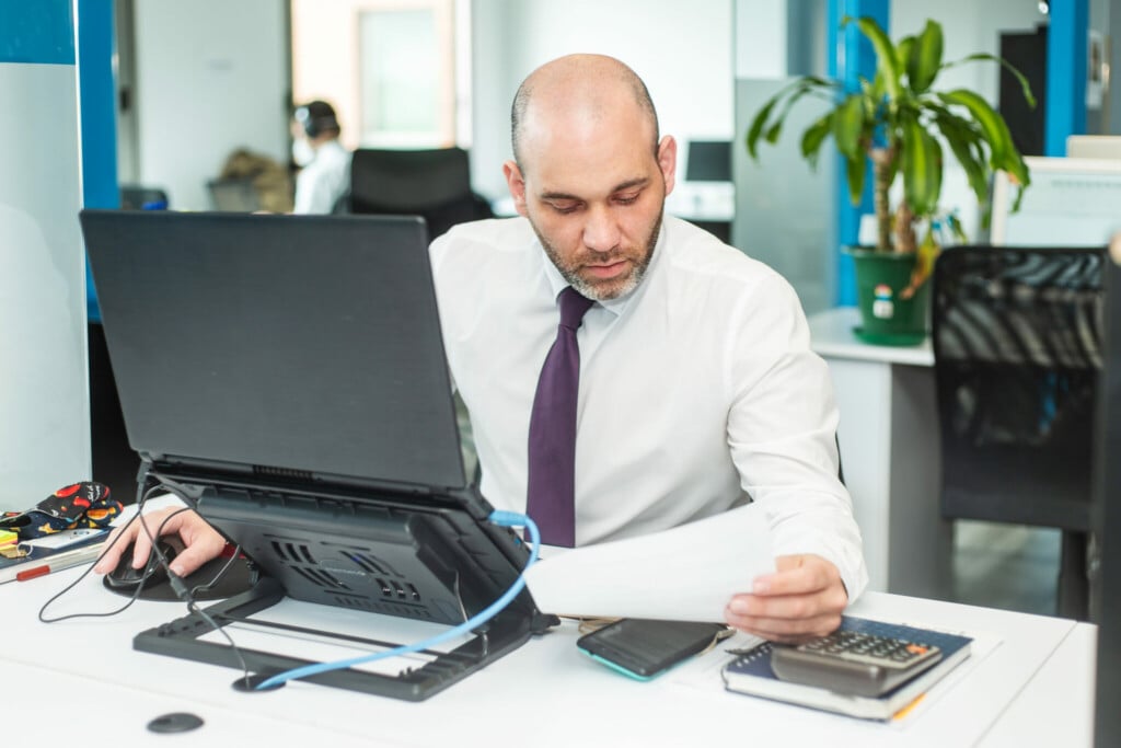 A man with a bald head and beard, wearing a white shirt and purple tie, sits at a desk in an office. He is intently looking at a sheet of paper in his right hand while using a computer mouse with his left. As this lawyer in Panama works, he is surrounded by a laptop, phone, and notebooks on the desk.