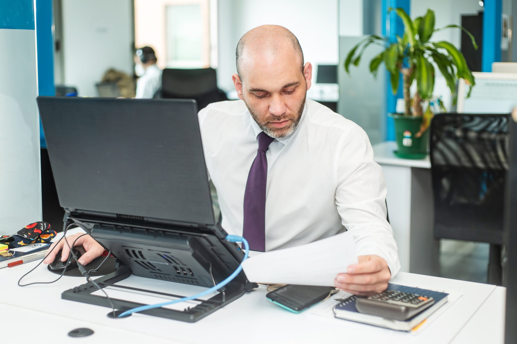 Um homem careca e barbudo, vestindo camisa branca e gravata roxa, senta-se à mesa de um escritório. Ele está olhando atentamente para uma folha de papel com a mão direita enquanto usa um mouse de computador com a esquerda. Enquanto esse advogado no Panamá trabalha, ele está cercado por um laptop, um telefone e notebooks sobre a mesa.  