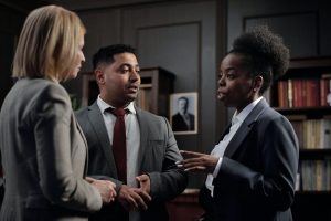 Three people in business attire are engaged in a conversation in an office setting. A woman with curly hair in a dark suit is speaking, while a man, who is an advogado no Brasil, in a gray suit with a red tie and a woman with blond hair in a light-colored suit listen attentively. Bookshelves are in the background.