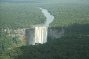 Vista aérea das Cataratas de Kaieteur, na Guiana, com água caindo em cascata de um penhasco em um desfiladeiro cercado por uma floresta tropical densa e exuberante. Um único rio serpenteia para longe da cachoeira através da floresta verde. O céu está parcialmente nublado, criando sombras esparsas. Os hotéis próximos a você na Guiana prometem uma estadia inesquecível.