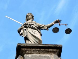 Una estatua de Lady Justice sosteniendo una espada en su mano derecha y una balanza en su izquierda se alza contra un cielo azul claro. La estatua, que simboliza la justicia y el derecho laboral en El Salvador, se encuentra sobre un pedestal de piedra. Los ojos de Lady Justice no están cubiertos por una venda.