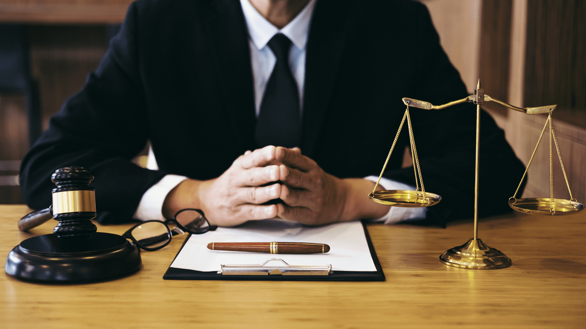 An attorney in El Salvador, dressed in a suit, sits at a desk with hands clasped. On the desk are a judge's gavel, glasses, a pen on a clipboard, and a balanced scale of justice. The background features shelves and wood paneling, suggesting a professional office or courtroom setting.