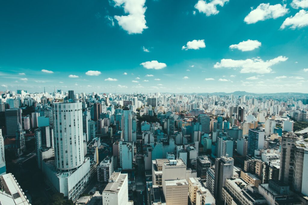 Aerial view of a large cityscape with numerous high-rise buildings under a partly cloudy sky. The foreground features tall, densely packed structures, while the background shows urban sprawl extending towards distant hills, evoking the meticulous search of caçadores de cabeças na Colômbia.