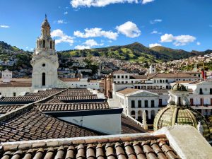 A cityscape view of Quito, Ecuador showing colonial-style buildings with tiled rooftops, encapsulating the charm and vibrancy of serviços back office no Equador. A white church with a bell tower stands prominently in the foreground. Hills covered in greenery and scattered houses are visible in the background under a partly cloudy sky.