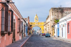 Uma rua de paralelepípedos ladeada por prédios coloridos leva a um arco amarelo com um relógio, conhecido como Arco de Santa Catalina, em Antígua, Guatemala. O caminho panorâmico, perfeito para um passeio depois que o senhor registra uma empresa na Guatemala, é cercado por vegetação, com poucos carros e pessoas visíveis.