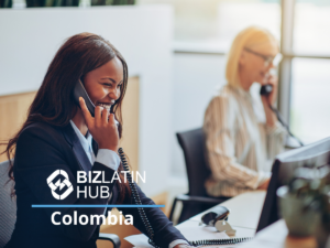 Two women are seated at desks in an office setting, both engaged in phone conversations. The woman in the foreground is smiling and appears to be speaking animatedly. The image features the logo and text "BizLatin Hub Colombia.