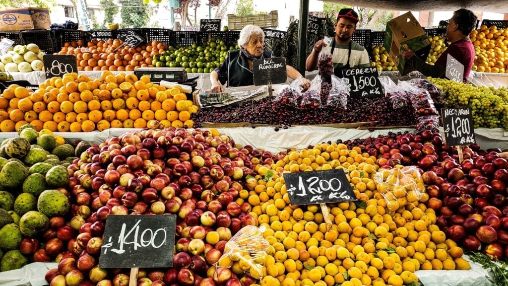 A market stall in Guatemala displays an assortment of fruits including apples, cherries, and peaches, with prices written on black signs. Three people behind the stall are doing business, one holding a box and interacting with each other. Various colors and quantities of fruits are visible.